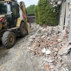 Żółta koparko-ładowarka pracuje przy rozbiórce starego domu. Gruz i cegły leżą na ziemi.
