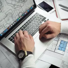 Rzeczoznawca z banku PKO analizuje plany architektoniczne na laptopie. Na biurku leży też telefon i długopisy.