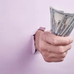 Dłoń przebija się przez różową ścianę, trzymając plik banknotów 100 USD. Może to symbolizować trudności z wynajmem bez umowy a wyprowadzką.