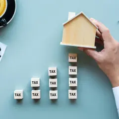 Domyślnie czy podatek od nieruchomości płaci się co roku? Drewniany domek na kostkach z napisem "TAX" symbolizuje roczne obciążenia.