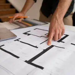 Ręce wskazujące na plany domu, obok laptopa i próbek materiałów.