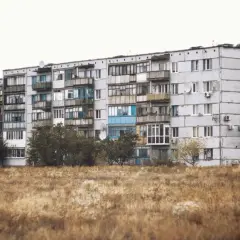 Szare bloki z balkonami, typowe jak wyglądają mieszkania socjalne. Przed nimi sucha trawa i drzewa.
