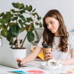 Młoda kobieta z kawą w ręku pracuje przy laptopie, zastanawiając się, ile trzeba pracować żeby dostać kredyt.
