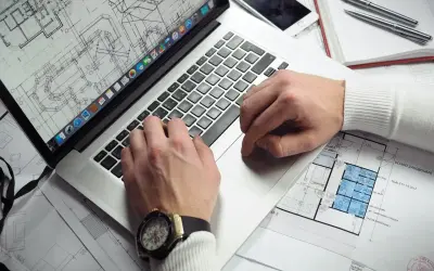 Rzeczoznawca z banku PKO analizuje plany architektoniczne na laptopie. Na biurku leży też telefon i długopisy.