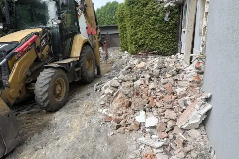 Żółta koparko-ładowarka pracuje przy rozbiórce starego domu. Gruz i cegły leżą na ziemi.