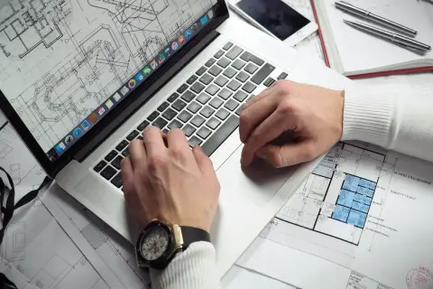 Rzeczoznawca z banku PKO analizuje plany architektoniczne na laptopie. Na biurku leży też telefon i długopisy.