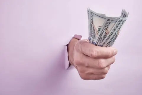 Dłoń przebija się przez różową ścianę, trzymając plik banknotów 100 USD. Może to symbolizować trudności z wynajmem bez umowy a wyprowadzką.