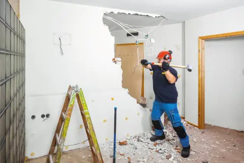 Mężczyzna w kasku i ochronnych słuchawkach rozbija ścianę kilofem. Widać przewody elektryczne i gruz. Zastanawiasz się, ile kosztuje wyburzenie ściany?