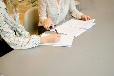Dwie kobiety przy stole omawiają dokumenty. Jedna pisze w notatniku, druga przegląda papiery. To może być umowa przyrzeczona co to znaczy.