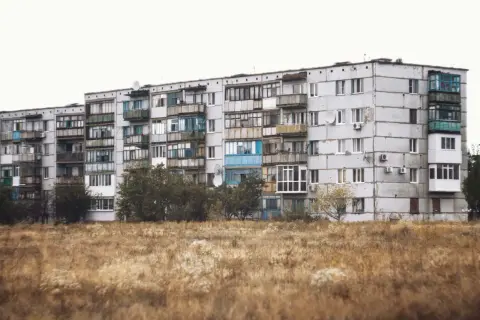 Szare bloki z balkonami, typowe jak wyglądają mieszkania socjalne. Przed nimi sucha trawa i drzewa.