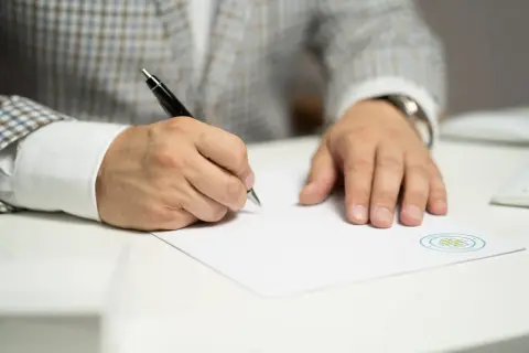 Mężczyzna w eleganckim garniturze pisze piórem na białej kartce, składając wniosek o wydanie listu mazalnego.