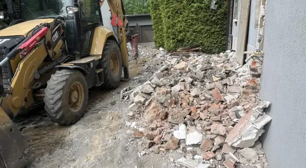 Żółta koparko-ładowarka pracuje przy rozbiórce starego domu. Gruz i cegły leżą na ziemi.