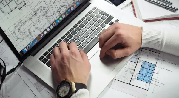 Rzeczoznawca z banku PKO analizuje plany architektoniczne na laptopie. Na biurku leży też telefon i długopisy.
