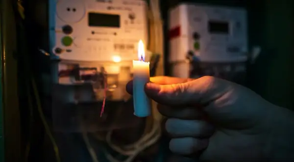 Płonąca świeca w dłoni na tle liczników energii elektrycznej. Gdy najemca nie płaci czynszu, można odłączyć mu media.