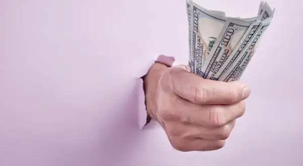 Dłoń przebija się przez różową ścianę, trzymając plik banknotów 100 USD. Może to symbolizować trudności z wynajmem bez umowy a wyprowadzką.