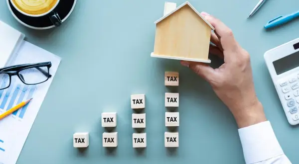 Domyślnie czy podatek od nieruchomości płaci się co roku? Drewniany domek na kostkach z napisem "TAX" symbolizuje roczne obciążenia.