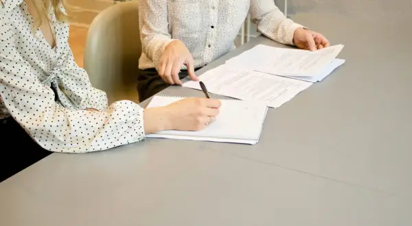 Dwie kobiety przy stole omawiają dokumenty. Jedna pisze w notatniku, druga przegląda papiery. To może być umowa przyrzeczona co to znaczy.