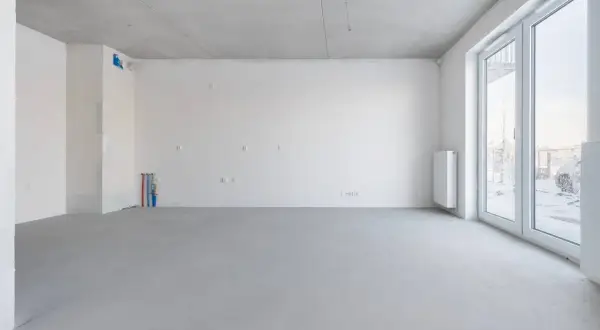 Pusta przestrzeń w stanie deweloperskim: betonowa posadzka, białe ściany, instalacje wodne i elektryczne, grzejnik, duże okna.