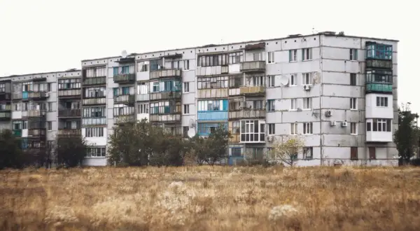 Szare bloki z balkonami, typowe jak wyglądają mieszkania socjalne. Przed nimi sucha trawa i drzewa.
