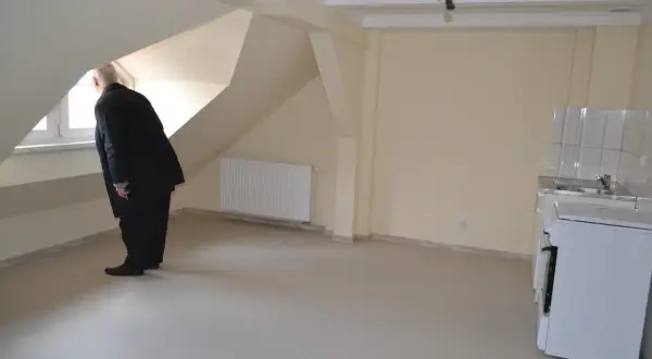 Starszy pan w garniturze ogląda puste mieszkanie, zastanawiając się, jak starać się o mieszkanie socjalne. Widać okno, kaloryfer i aneks kuchenny.