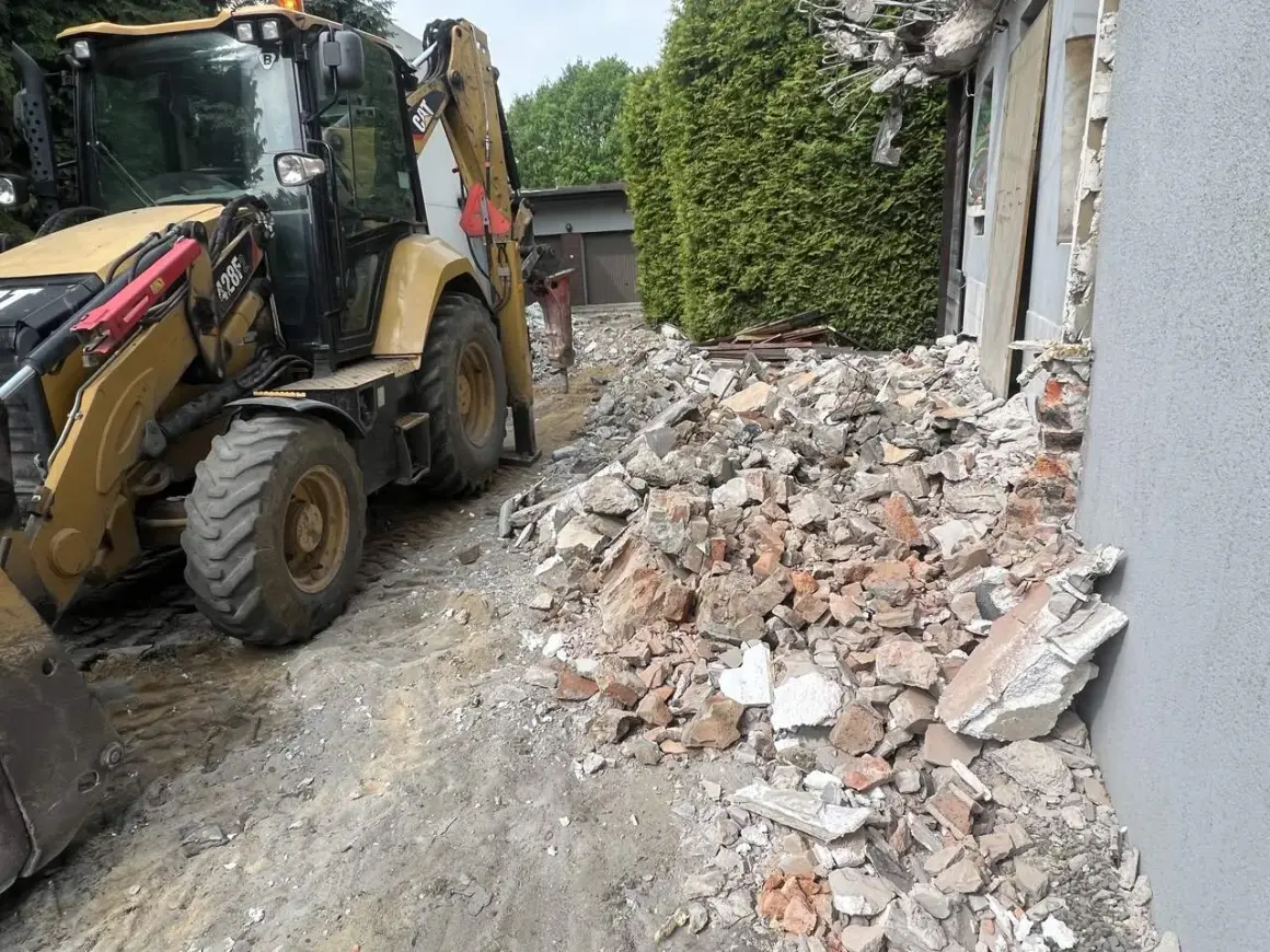 Żółta koparko-ładowarka pracuje przy rozbiórce starego domu. Gruz i cegły leżą na ziemi.