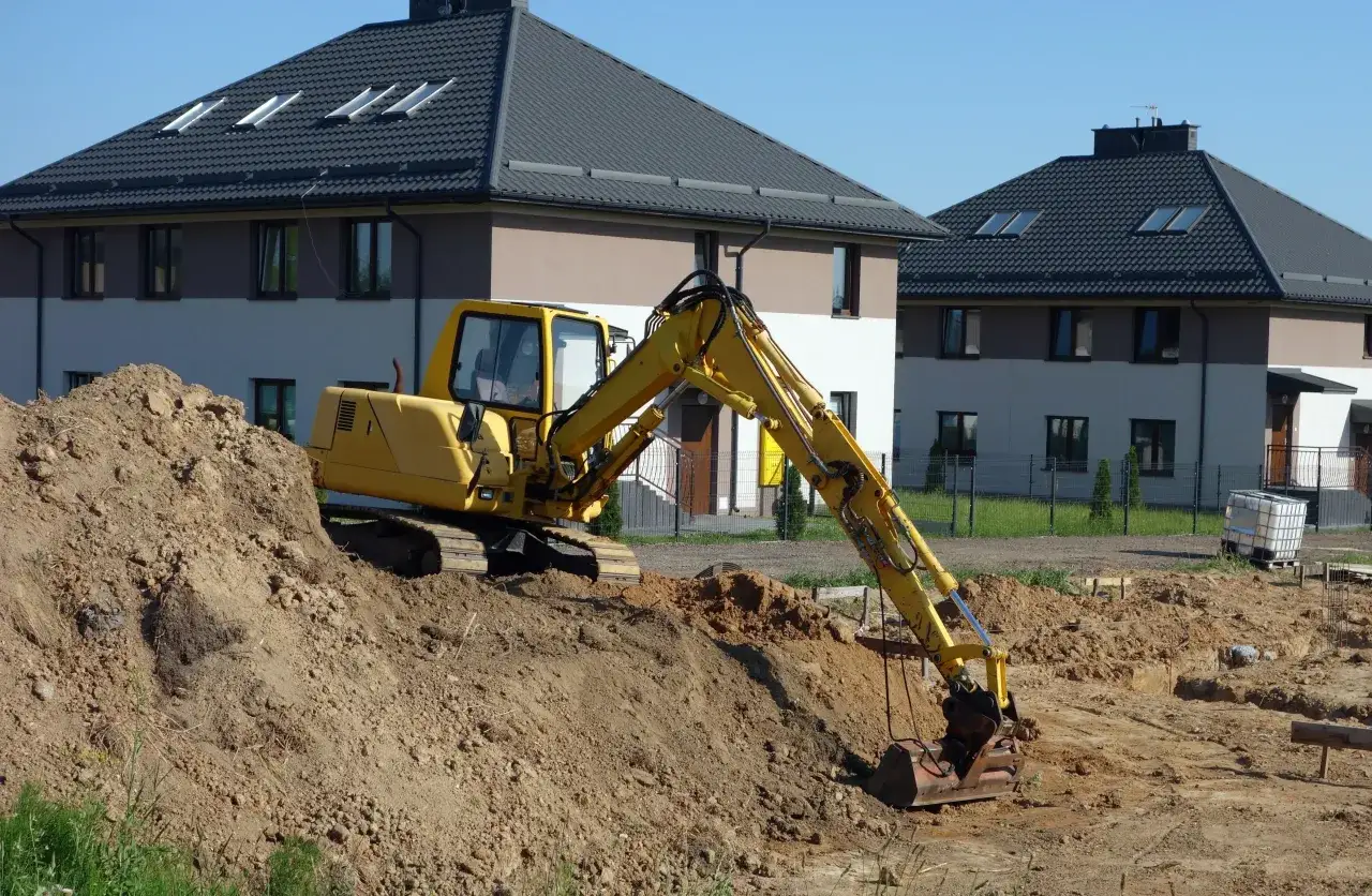 Żółta koparka pracuje na budowie, przygotowując teren pod budowę domu. To pierwszy krok do własnego lokum, zamiast mieszkania na start.