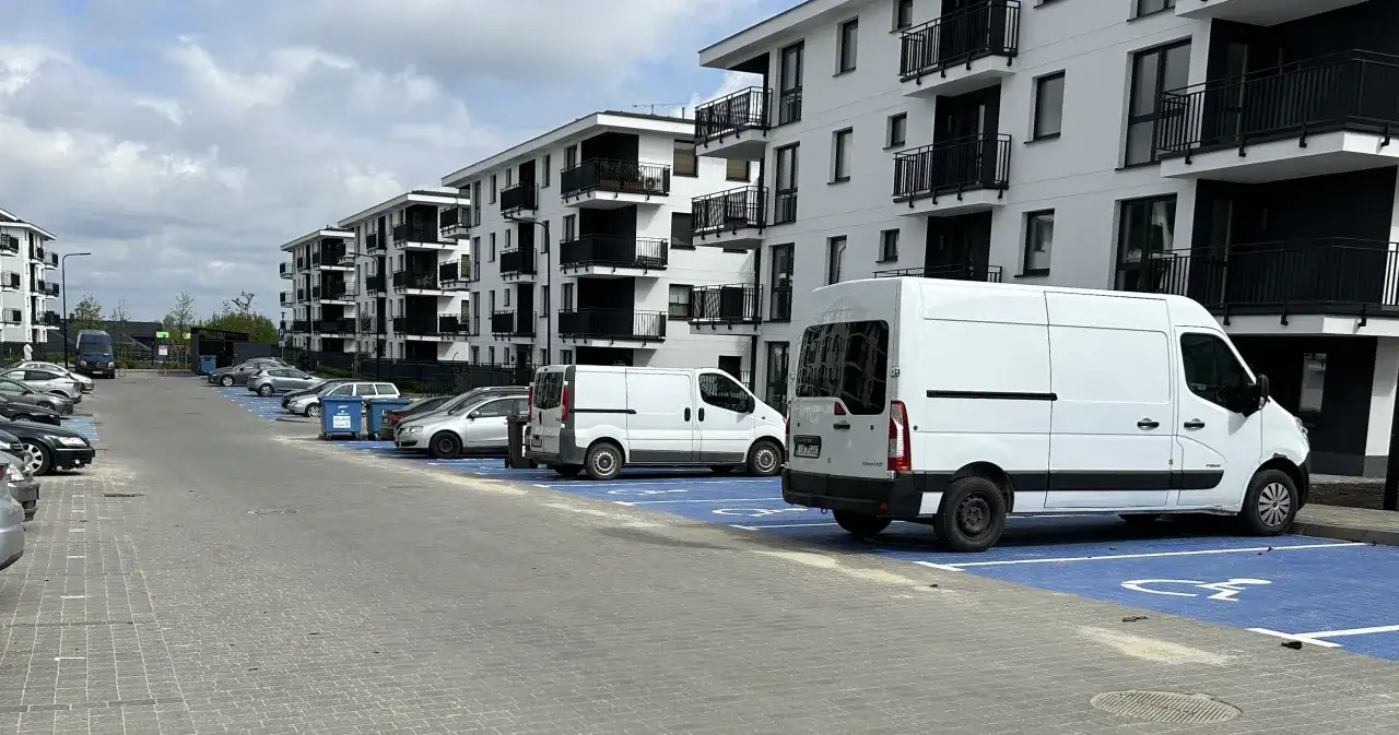 Nowoczesne osiedle z białymi blokami i czarnymi balkonami. Na parkingu, z niebieskimi miejscami, stoi kilka samochodów i białe busy. Zastanawiamy się, ile miejsc parkingowych na mieszkanie przewidziano.