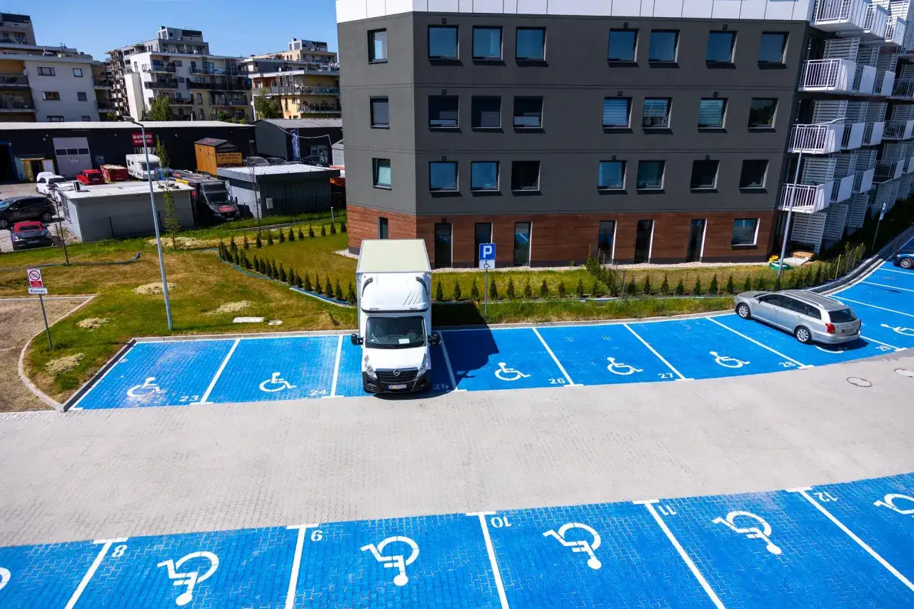 Parking dla osób niepełnosprawnych z niebieskimi oznaczeniami. Widać samochód dostawczy i osobowy. Ile miejsc parkingowych na mieszkanie?