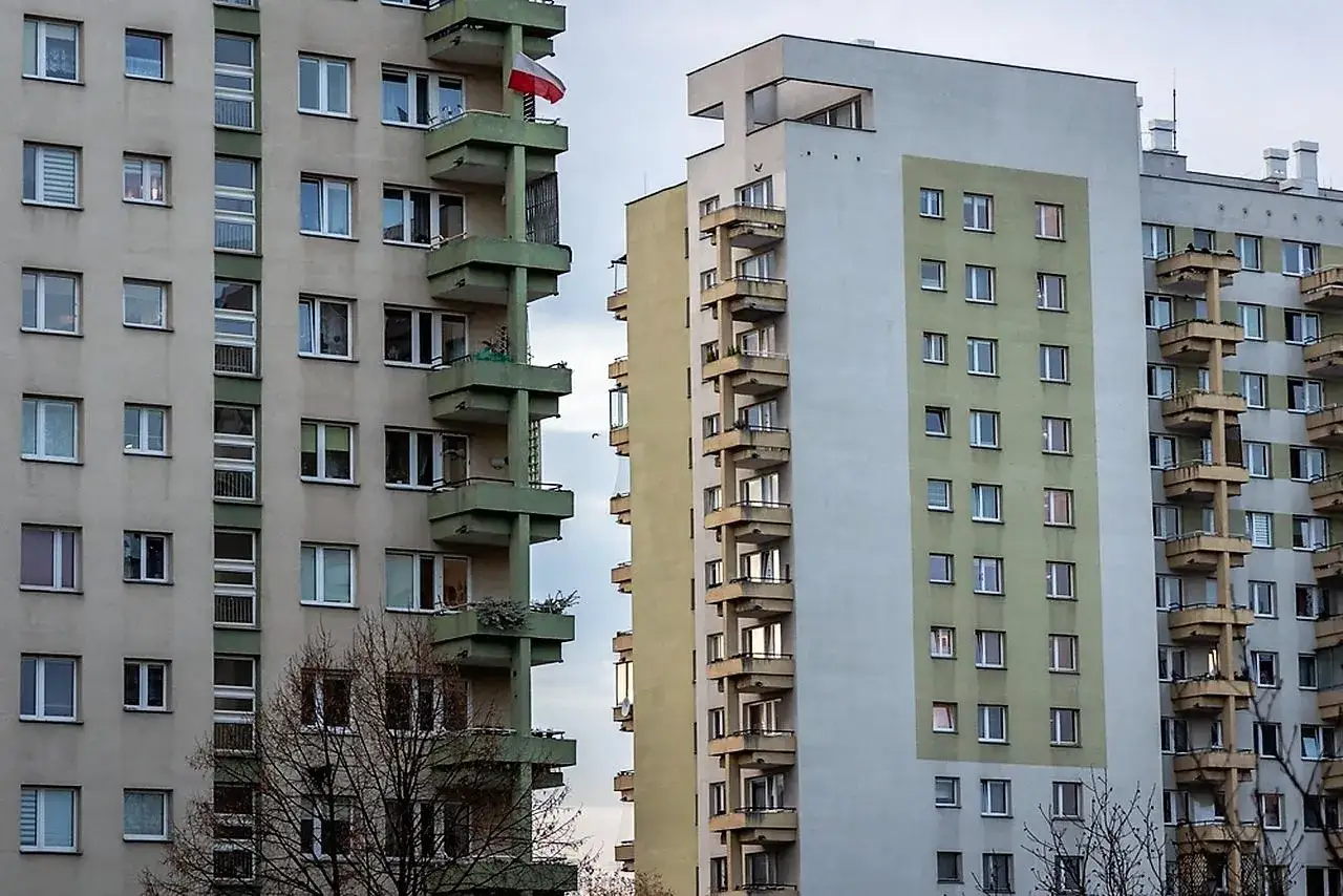 Wielopiętrowy dom z wielkiej płyty z zielonymi balkonami i polską flagą.