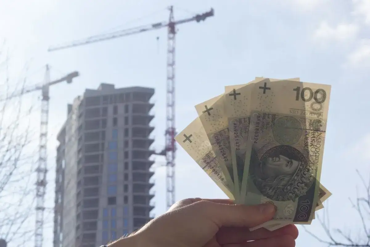 Dłoń trzyma polskie banknoty 100 zł na tle budowy wieżowca i dźwigów. Sytuacja na rynku nieruchomości jest dynamiczna.