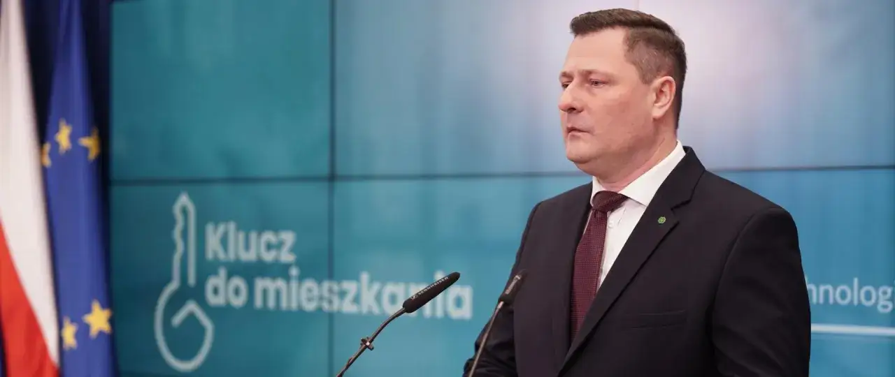 Mężczyzna w garniturze przemawia na tle logo programu mieszkaniowego 