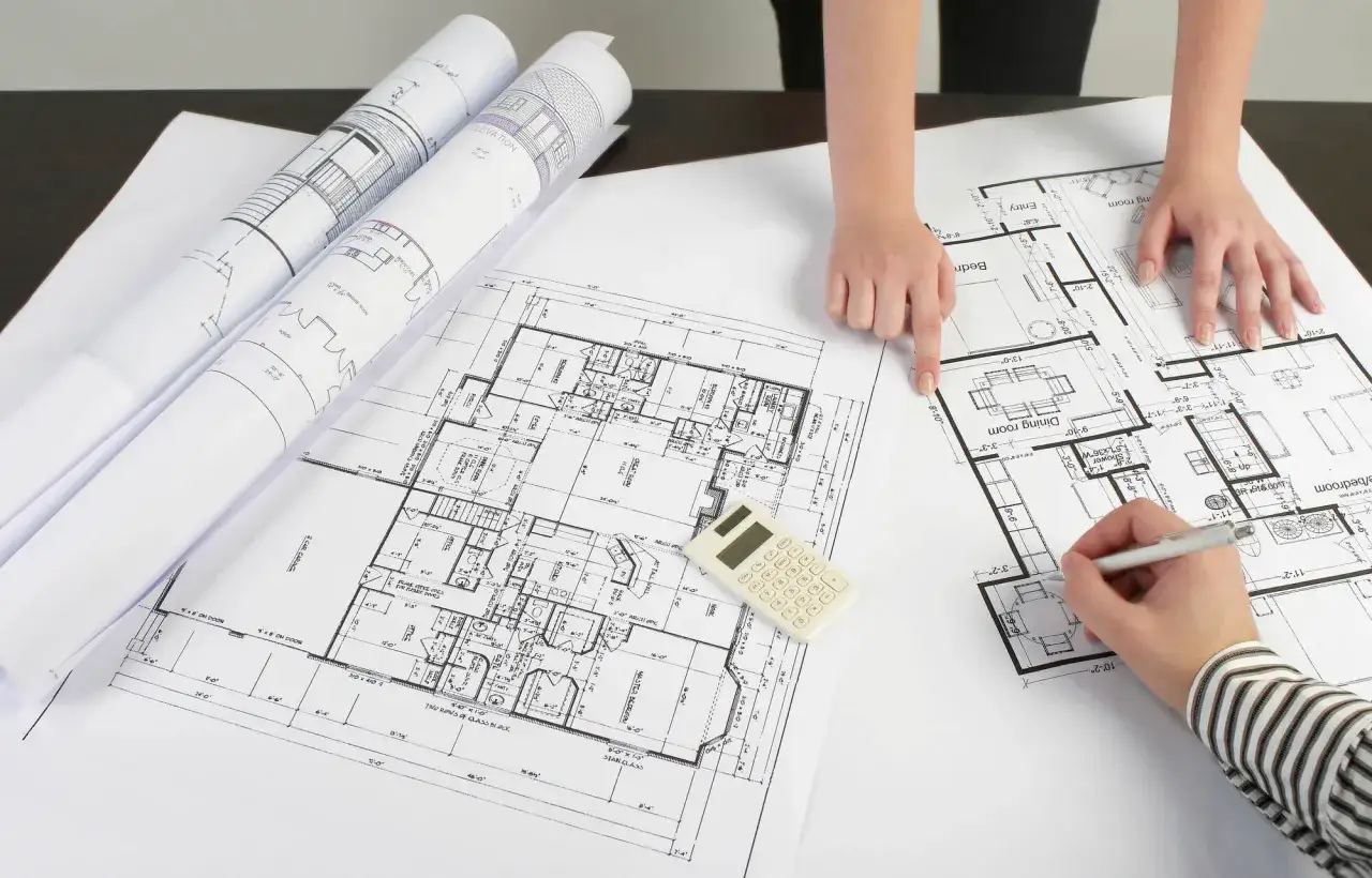 Dwie osoby analizują plany architektoniczne, dyskutując, czy adaptacja projektu jest konieczna. Na stole leżą zwinięte projekty i kalkulator.