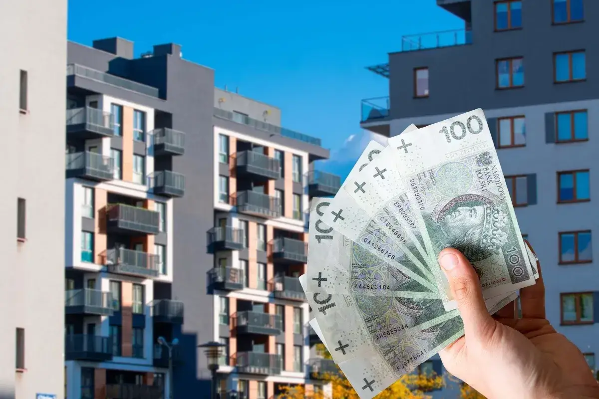 Dłoń trzyma banknoty 100 zł przed nowoczesnymi blokami mieszkalnymi. To może być zaliczka eksploatacyjna na nowe mieszkanie.