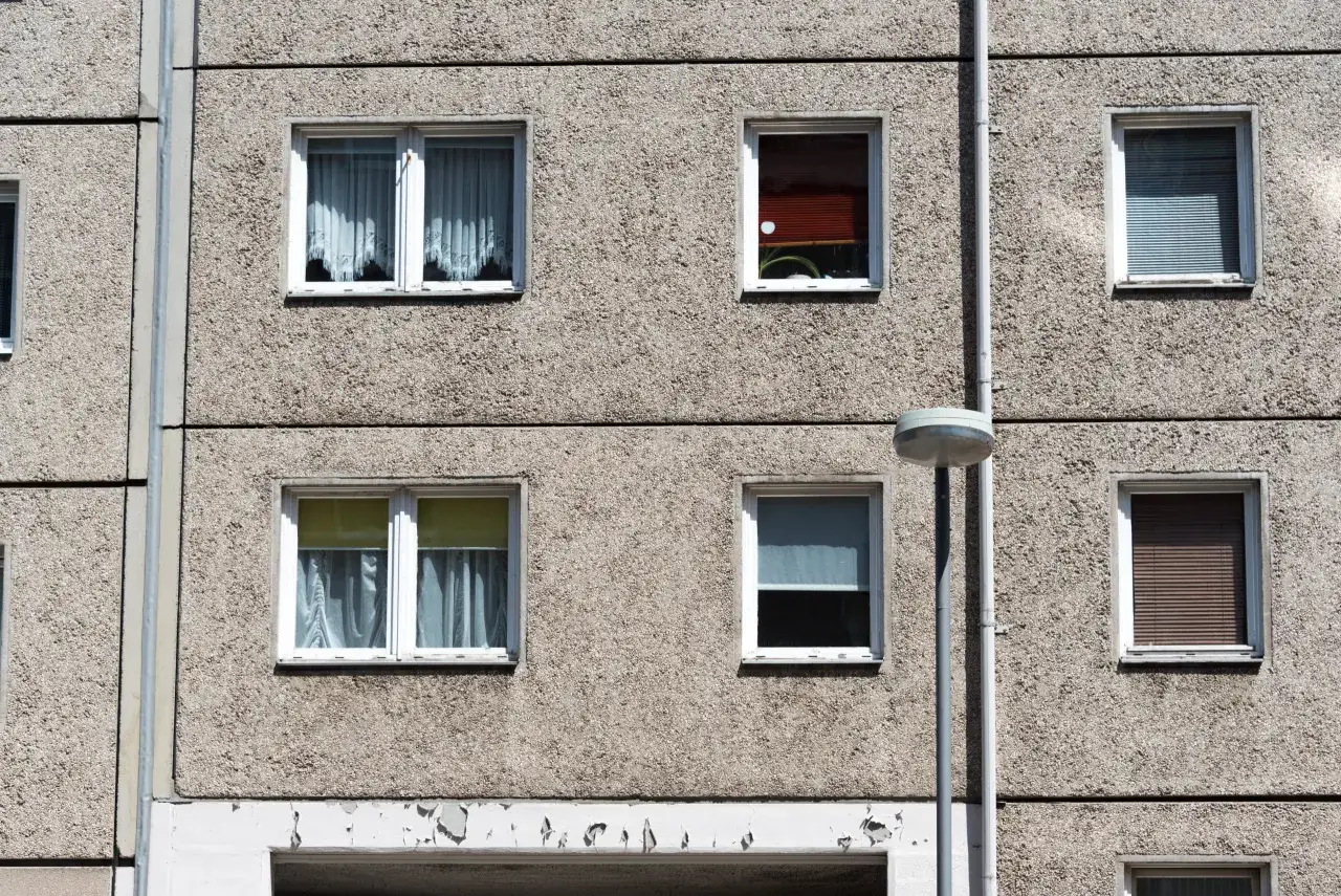 Ściana domu z wielkiej płyty z kilkoma oknami, niektóre z zasłonami, inne z roletami.