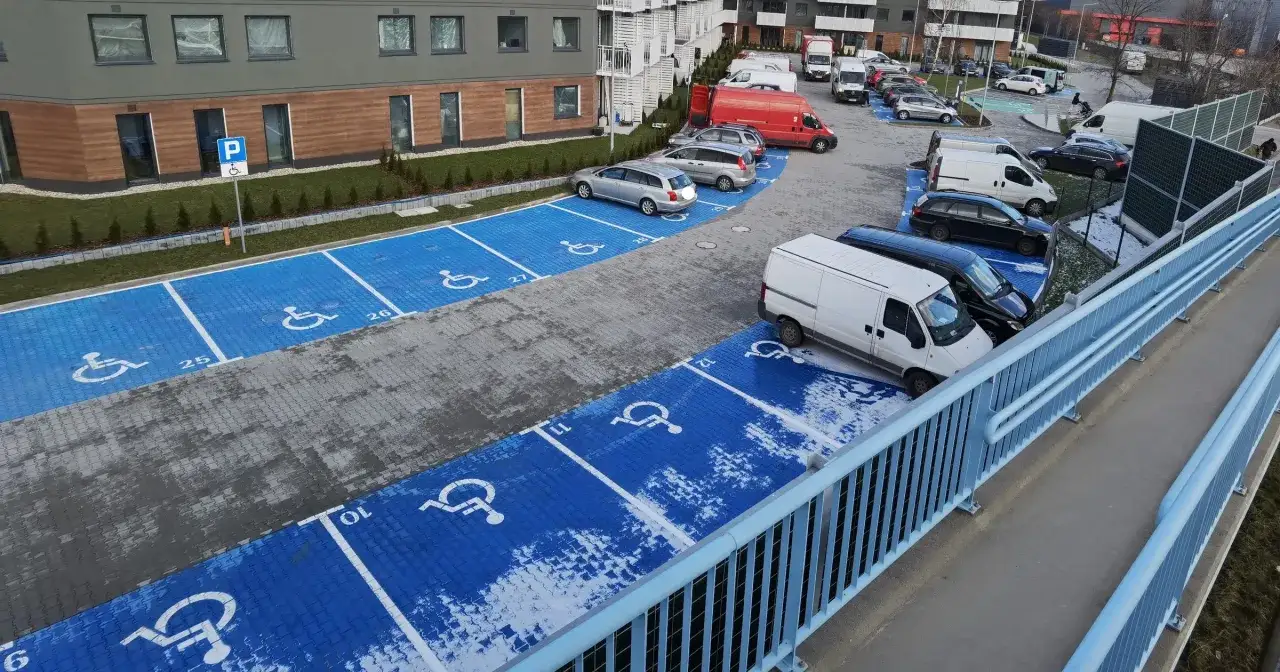Niebieskie miejsca parkingowe dla niepełnosprawnych z numerami, obok budynków mieszkalnych. Trudno ocenić ile miejsc parkingowych na mieszkanie.