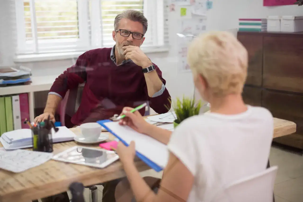 Rozmowa o tym, ile zarabia członek zarządu spółdzielni mieszkaniowej. Mężczyzna w okularach słucha kobiety notującej na kartce.