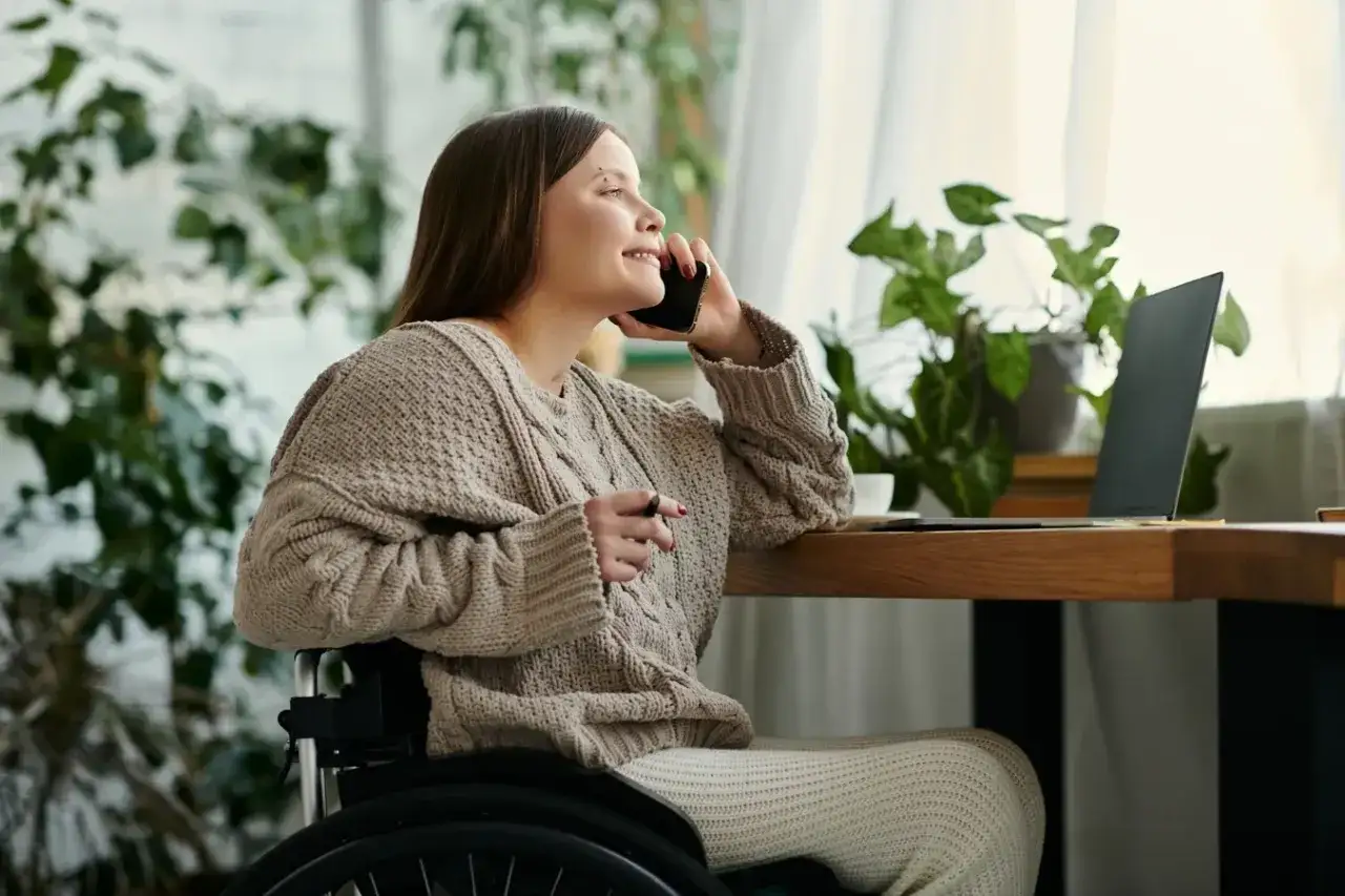 Kobieta na wózku inwalidzkim rozmawia przez telefon, planując dofinansowanie do zakupu mieszkania. Obok laptop i rośliny.