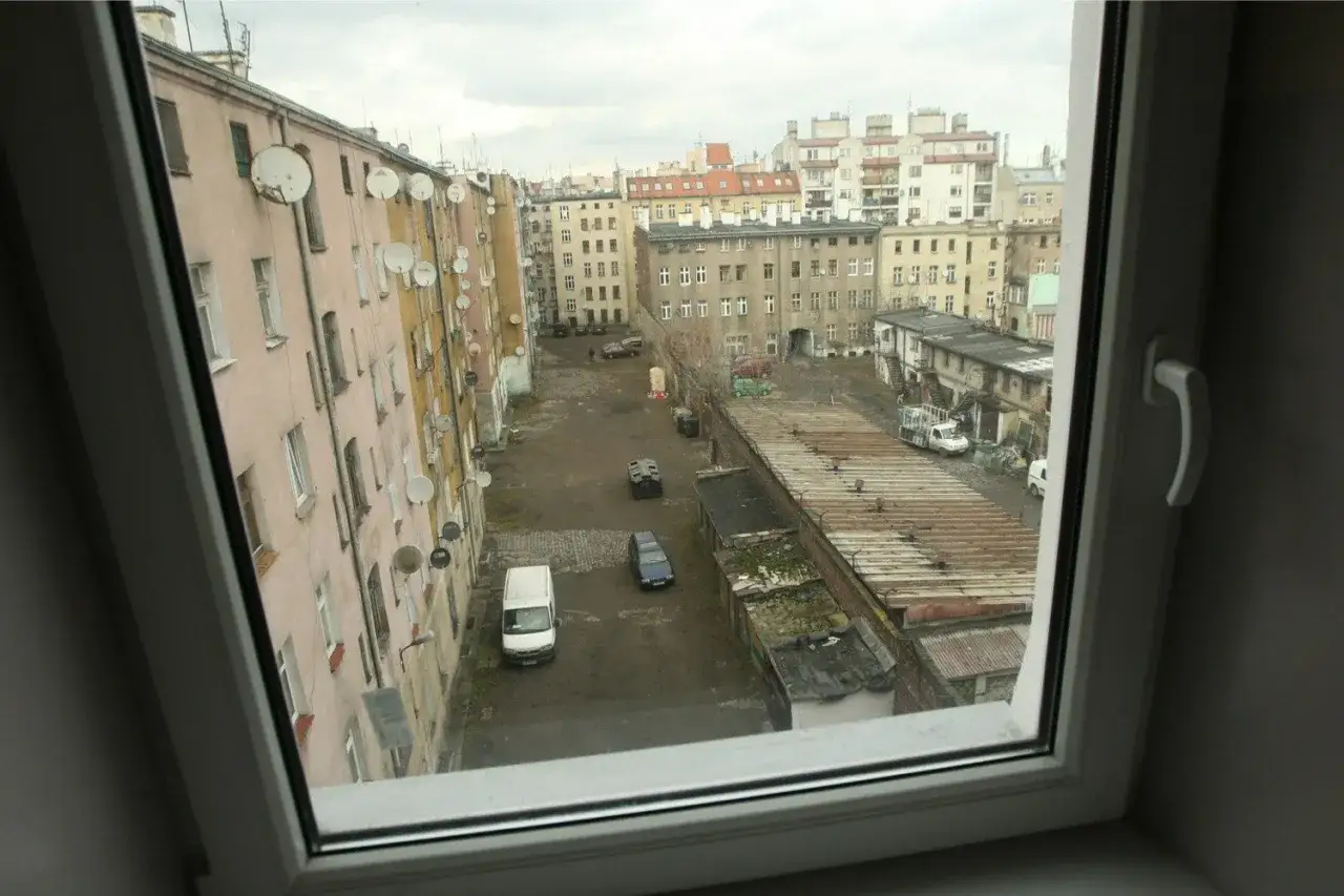 Podwórko kamienic, gdzie widać, jak wyglądają mieszkania socjalne: zaniedbane budynki, szare podwórko, samochody i anteny satelitarne.
