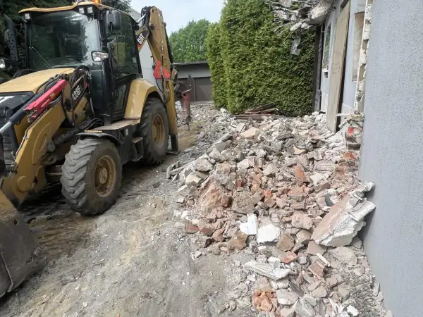 Żółta koparko-ładowarka pracuje przy rozbiórce starego domu. Gruz i cegły leżą na ziemi.