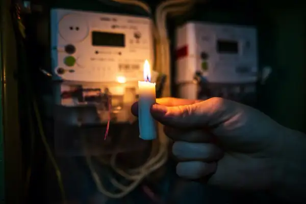 Płonąca świeca w dłoni na tle liczników energii elektrycznej. Gdy najemca nie płaci czynszu, można odłączyć mu media.